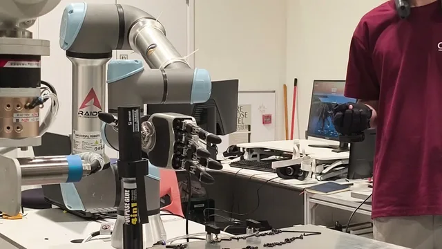 Teleoperated UR arm with a three-fingered hand applying glue on a workbench.