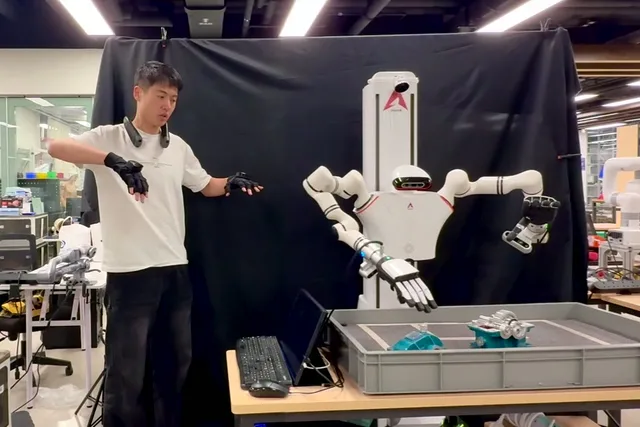 Operator in gloves and a neck-worn controller teleoperating a dual-arm robot over a parts tray.